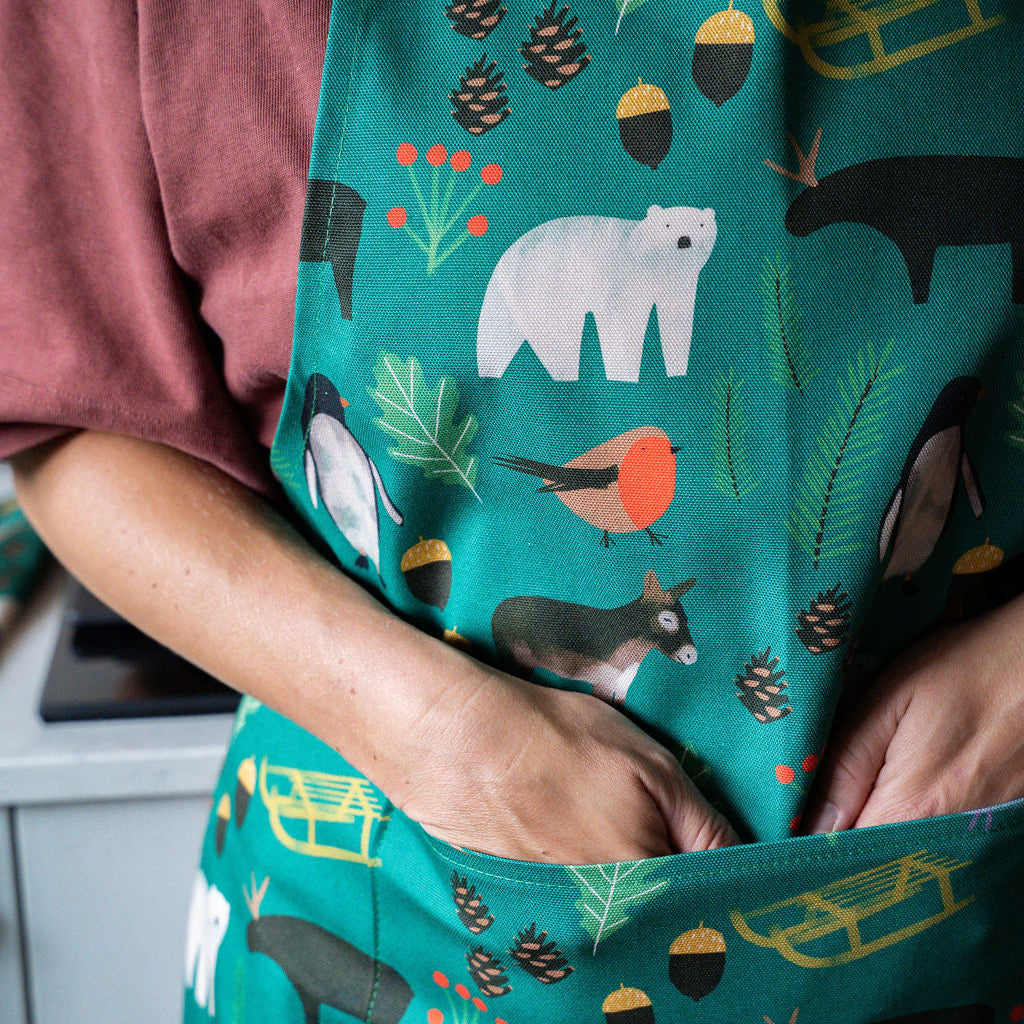 A close-up of a person wearing a green apron with a winter woodland print featuring polar bears, penguins, a red-breasted bird, pine cones, acorns, and sleighs. The person's hands are tucked into the front pocket.
