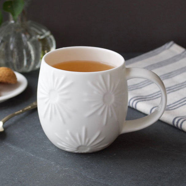 White embossed mug with tea on a dark surface with cookies and a vase in the background