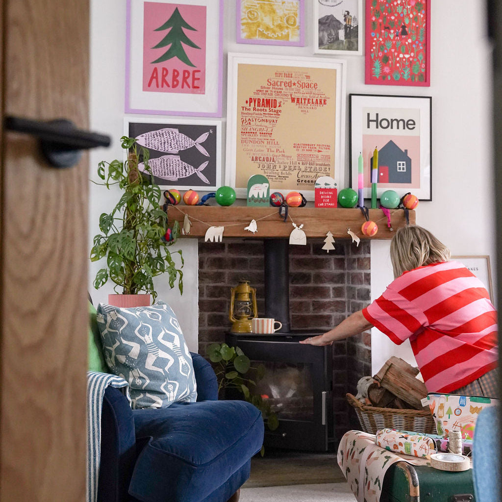 A cosy living room scene with a woman in a red and white striped shirt standing by a brick fireplace. The wall above the fireplace is decorated with a colorful collection of framed art prints.