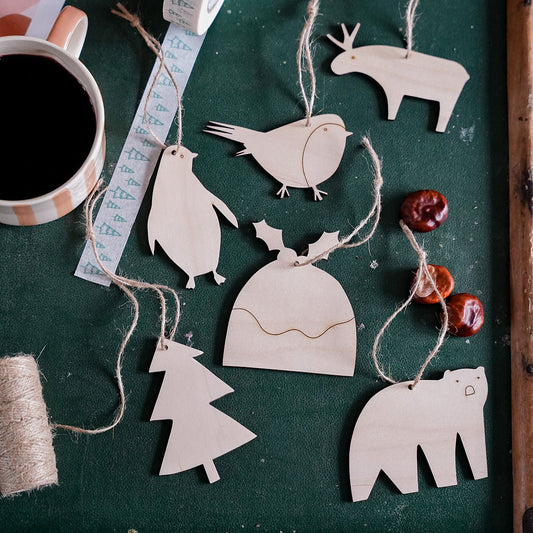 A flat-lay of natural wood Christmas ornaments shaped like a reindeer, penguin, bird, Christmas tree, Santa hat, polar bear, and holly, hanging on a dark green suitcase next to a cup of black coffee and holiday-themed tape.