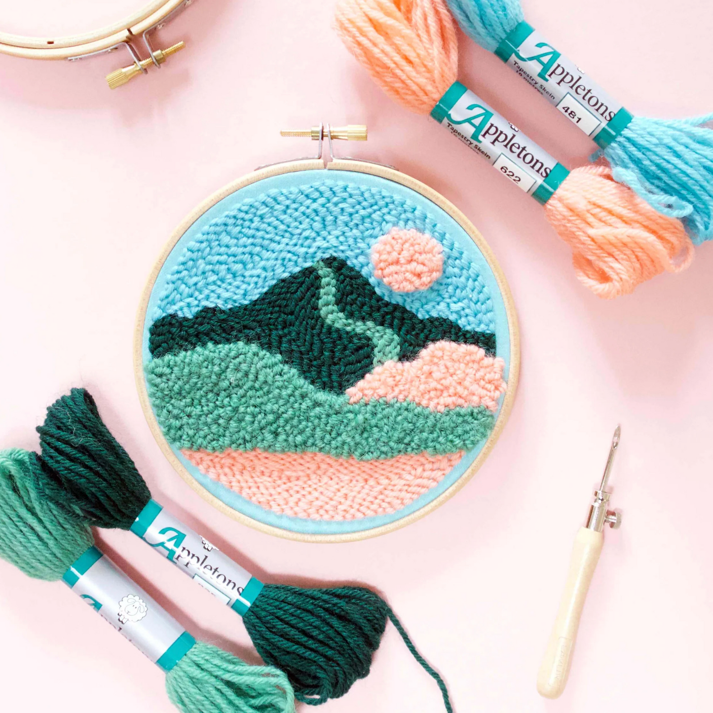 Embroidery hoop with mountain design, Appletons yarn, and needle on a pink background