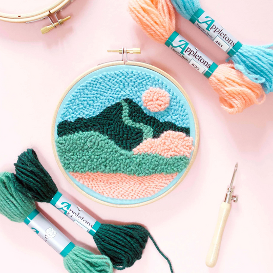 Embroidery hoop with mountain design, Appletons yarn, and needle on a pink background