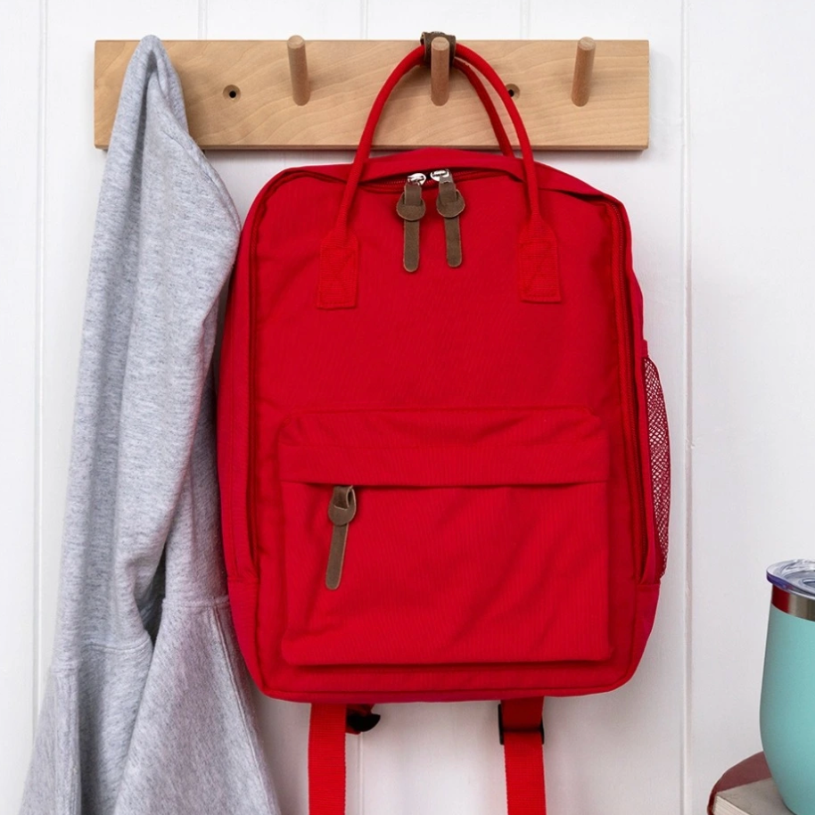 A bright red backpack with dual top handles and a front zip pocket hangs on a wooden peg rack against a white paneled wall. A gray hoodie is hanging to the left, and a small portion of a light blue tumbler is visible at the lower right.
