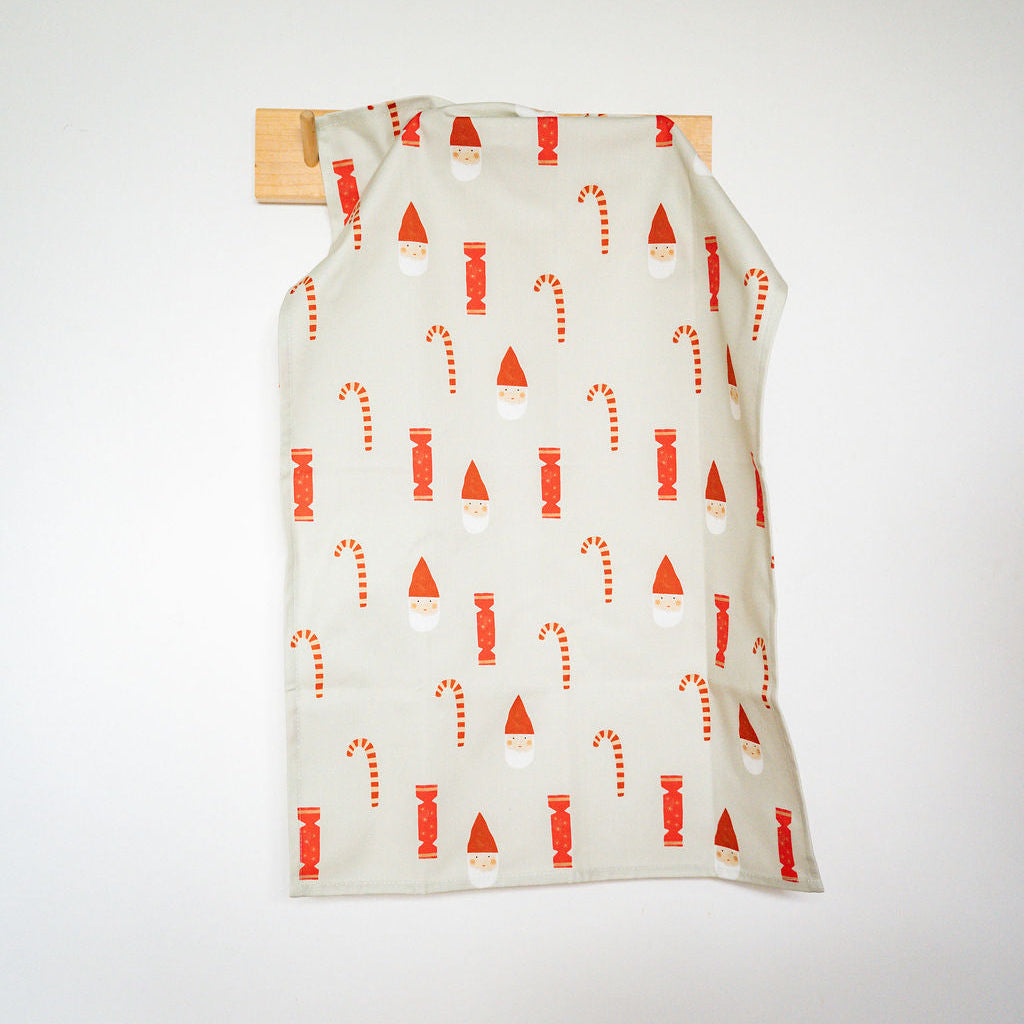 A cream-coloured fabric garment or towel with a red and white holiday pattern of Santa hats and candy canes, hanging from a wooden peg on a white wall.