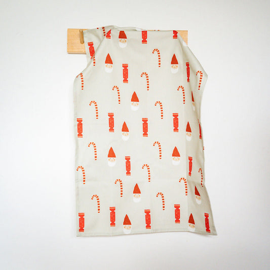 A cream-coloured fabric garment or towel with a red and white holiday pattern of Santa hats and candy canes, hanging from a wooden peg on a white wall.