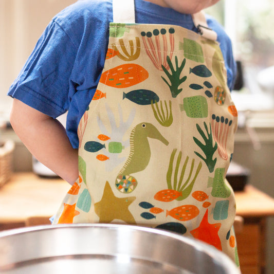 A child wearing a beige apron with colorful sea creature patterns, 100% cotton, adjustable strap. Made in Britain.