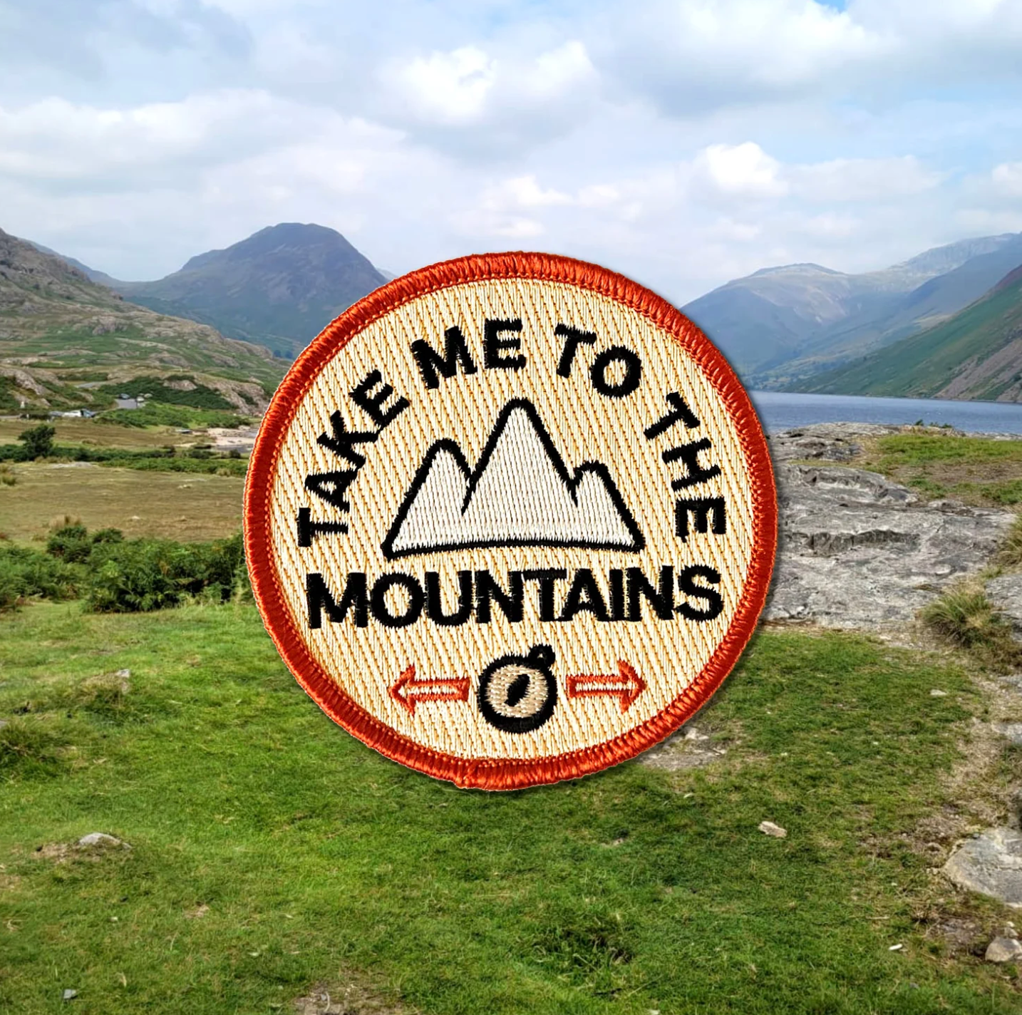Round embroidered patch with 'Take Me to the Mountains' text and mountain graphic, set against a scenic mountain landscape.