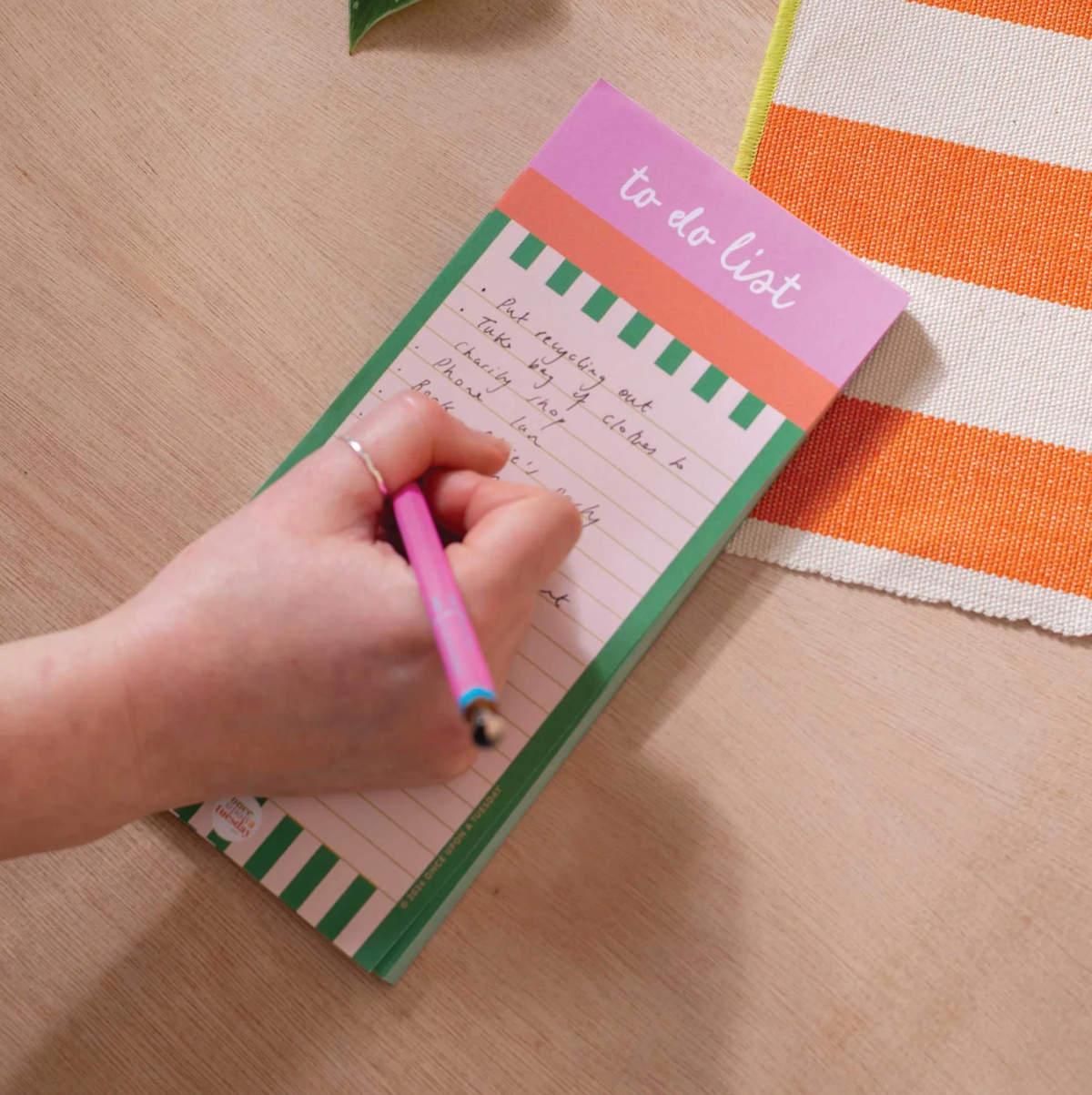 A hand holding a pink pen writes on a pastel to-do list notepad with green and orange striped borders, placed on a light wood tabletop next to an orange and white striped fabric.