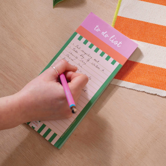 A hand holding a pink pen writes on a pastel to-do list notepad with green and orange striped borders, placed on a light wood tabletop next to an orange and white striped fabric.