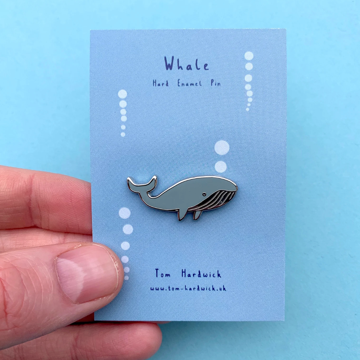Whale enamel pin held by a hand on a blue card with branding details.