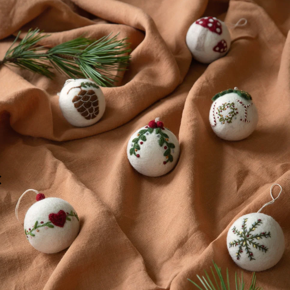Six white felt Christmas ornaments with hand-painted holiday designs, including a pinecone, mistletoe, holly, snowflake, and red berries, arranged on a draped tan fabric with pine branches.