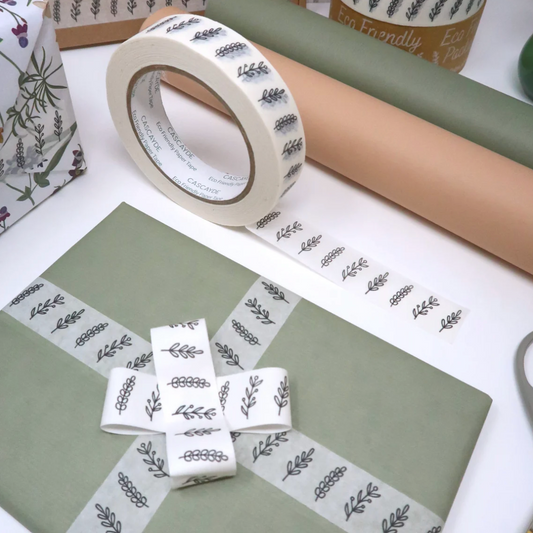 A roll of decorative washi tape with a repeating gray botanical leaf pattern on a white background, shown alongside wrapped gift boxes in muted green and tan tones on a white tabletop.