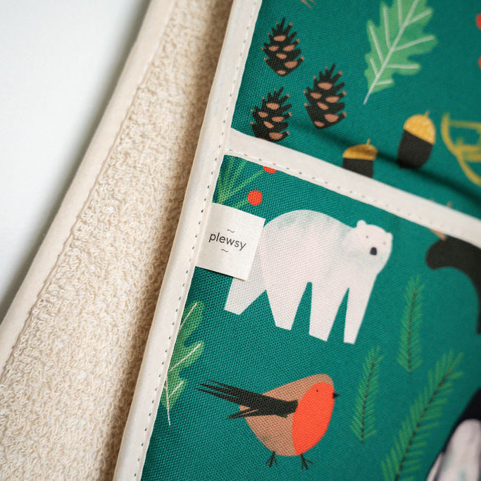 A close-up of plewsy double oven gloves, featuring a green winter themed print with a polar bear, bird, pinecones, and a birdhouse, paired with a beige textured lining. A small white tag reads 'plewsy'.