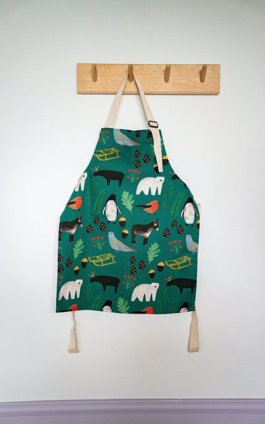 A green children's apron with a whimsical animal and nature print, hanging from a wooden peg on a white wall.