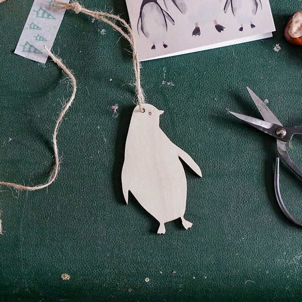 A styled flat-lay of a handmade penguin ornament on a dark green suitcase, surrounded by crafting supplies including scissors, twine, a coffee mug, and a penguin card reading 'Christmas Love'.