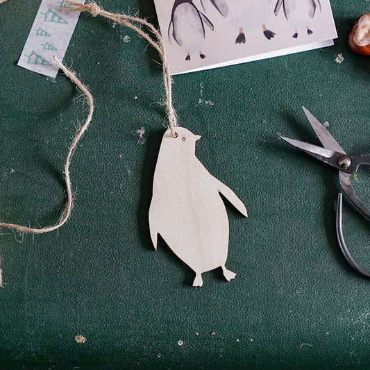 A styled flat-lay of a handmade penguin ornament on a dark green suitcase, surrounded by crafting supplies including scissors, twine, a coffee mug, and a penguin card reading 'Christmas Love'.