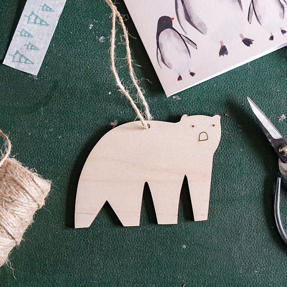 A styled flat-lay of Christmas crafting supplies on a dark green surface, including a wooden polar bear decoration, a penguin card reading 'Christmas Love', a spool of twine, scissors, and small decorative items.